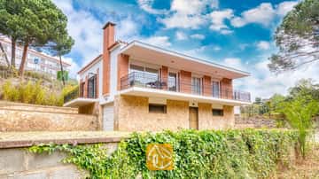 Außenansicht der Villa Lloret mit einem zweistöckigen Haus, einem Erdgeschoss aus Naturstein, einem Obergeschoss aus Backstein und einem umlaufenden Balkon unter einem wolkigen blauen Himmel.
