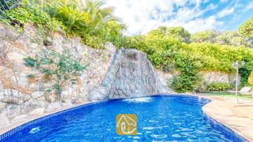 Ein sprudelnder Felsen-Wasserfall, der in einen blau gefliesten Swimmingpool der Villa Alba fließt, umgeben von Natursteinmauern und üppigem Grün unter einem leicht bewölkten Himmel.
