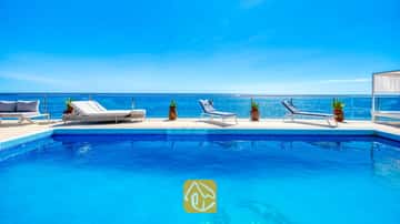 Der leuchtend blaue Swimmingpool der Villa Infinity mit weißen Sonnenliegen und Pflanzkübeln auf der gefliesten Poolterrasse mit Blick auf das Meer
