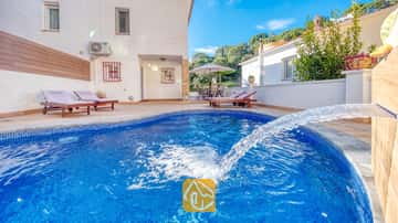 Geschwungener, blau gefliester Swimmingpool mit Wasserspiel in der Villa Blanca, ausgestattet mit hölzernen Sonnenliegen auf der gepflasterten Terrasse vor einer weißen Außenwand.