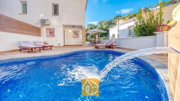 Geschwungener, blau gefliester Swimmingpool mit Wasserspiel in der Villa Blanca, ausgestattet mit hölzernen Sonnenliegen auf der gepflasterten Terrasse vor einer weißen Außenwand.