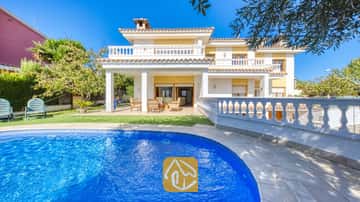 Ein geschwungener, blau gefliester Swimmingpool im Vordergrund, dahinter die Villa Baileys mit ihrer weißen Außenfassade, gelben Wänden und gefliesten Balkonen unter einem klaren Himmel.