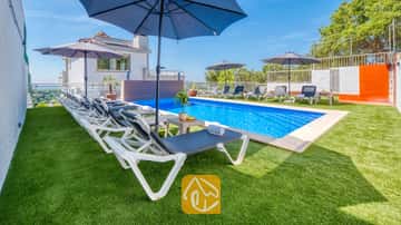Privater Pool in der Villa Sandy Beach mit einer Reihe weißer Sonnenliegen, blauen Sonnenschirmen und einem grünen Kunstrasen unter strahlend blauem Himmel.