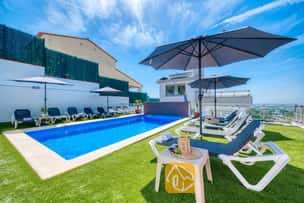 Privater Pool in der Villa Sandy Beach, umgeben von Kunstrasen, grauen Sonnenliegen mit weißem Rahmen und großen Sonnenschirmen unter einem strahlend blauen Himmel.