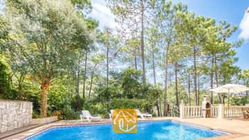 Der private Pool der Villa Esmee lädt mit weißen Sonnenliegen auf der gefliesten Terrasse, umgeben von hohen Pinien und einem Steingrill, zum Entspannen ein.