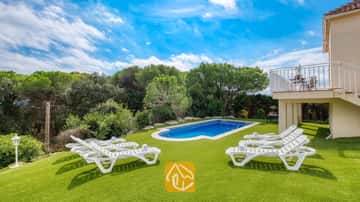 Außenpool, umgeben von einer grünen Wiese und weißen Sonnenliegen, in der Villa La Luna in Spanien, vor einer malerischen Kulisse aus Bäumen und blauem Himmel.