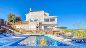 Ein rechteckiger Swimmingpool vor der Steinfassade der Villa Abigail unter einem klaren blauen Himmel, umgeben von Sonnenliegen und einem Sonnenschirm.