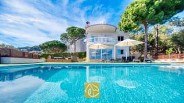 Die Außenanlage der Villa Chanel besticht durch einen türkisfarbenen Swimmingpool, weiße Sonnenschirme auf der Terrasse sowie einen geschwungenen Steinbalkon, der von üppigen grünen Pinien umgeben ist.