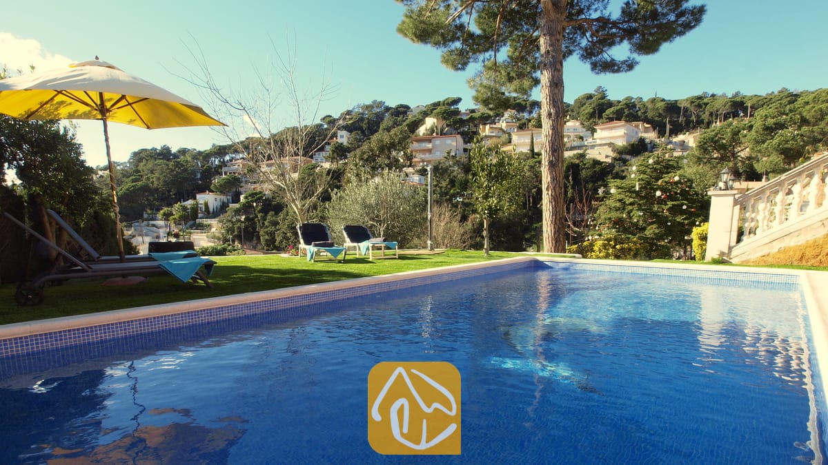 Ein privater Pool mit blau gefliestem Rand in der Villa Noa, ausgestattet mit Sonnenliegen auf der Wiese und Blick auf den Hang unter einem klaren Himmel.