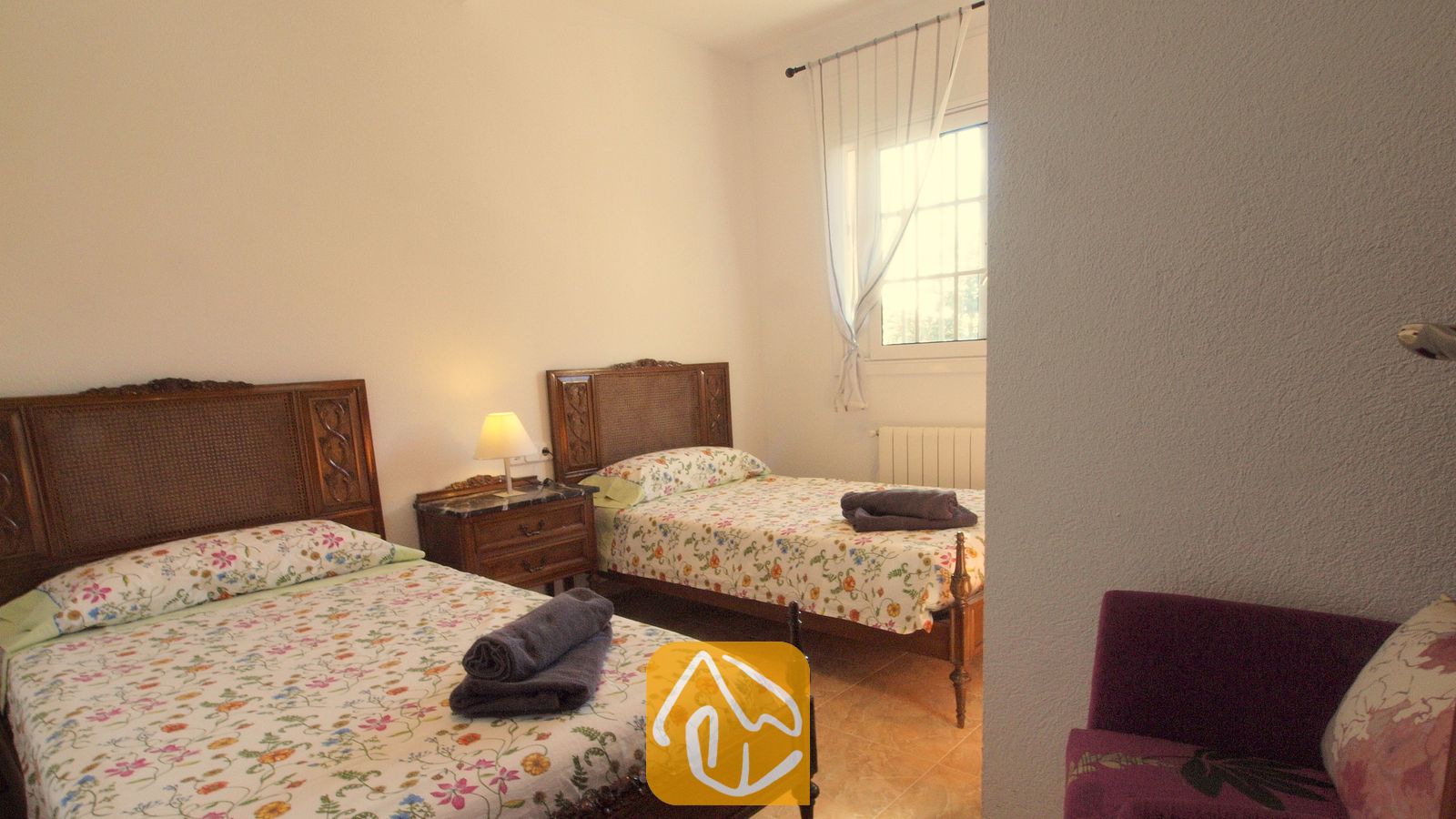 Schlafzimmer mit zwei Einzelbetten, dunklen Holzkopfteilen, mit Blumenmustern bedruckten Tagesdecken und gerollten grauen Handtüchern.