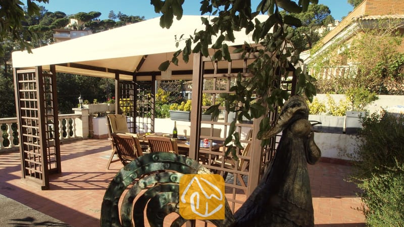 Holzpergola mit Stoffdach auf einer sonnigen Terrasse der Villa Noa, ausgestattet mit einem Esstisch im Freien und Stühlen im Schatten.