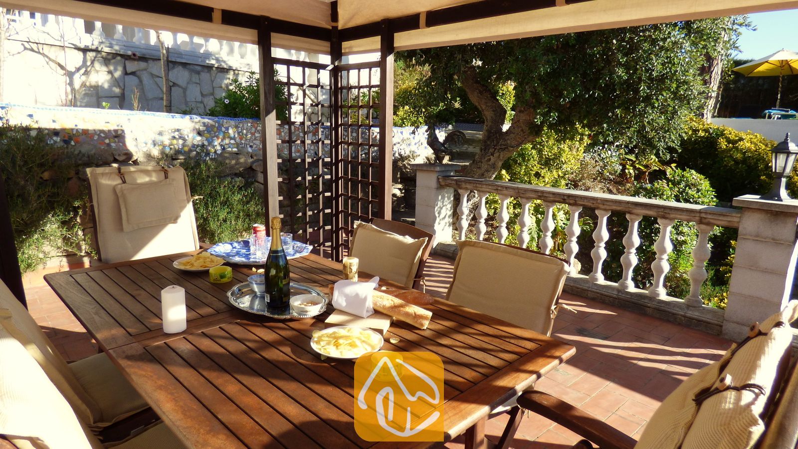 Überdachte Holz-Essterrasse der Villa Noa mit einem Tisch, der mit Wein, Snacks und Kerzen gedeckt ist, und Blick auf einen Garten mit steinerner Balustrade.