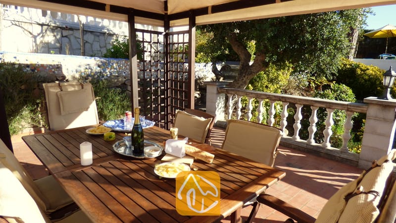Überdachte Holz-Essterrasse der Villa Noa mit einem Tisch, der mit Wein, Snacks und Kerzen gedeckt ist, und Blick auf einen Garten mit steinerner Balustrade.