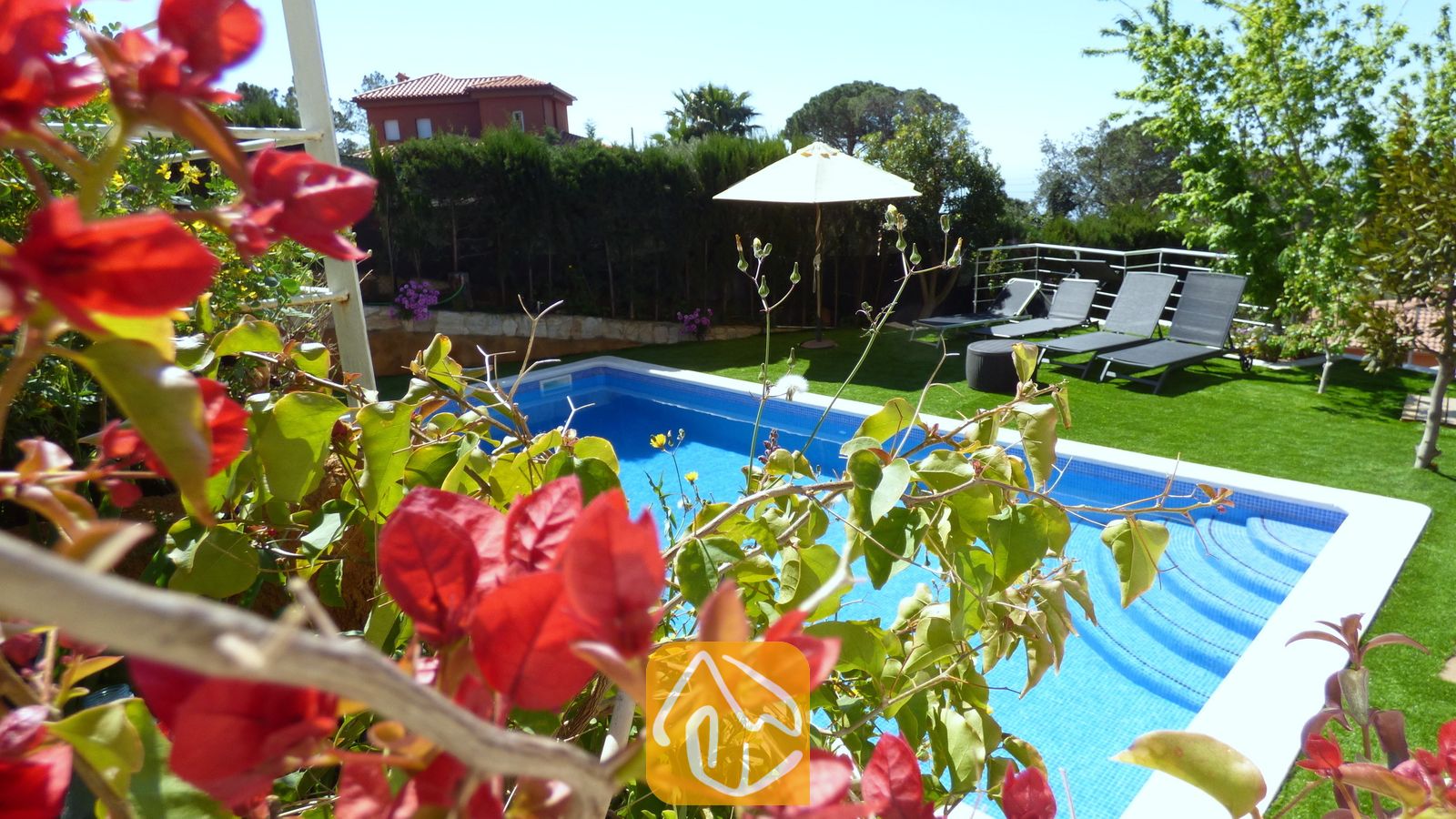 Der private Pool der Villa Noa, eingerahmt von roten Bougainvillea-Blüten, mit Sonnenliegen und einem weißen Sonnenschirm auf dem Rasen im Hintergrund.