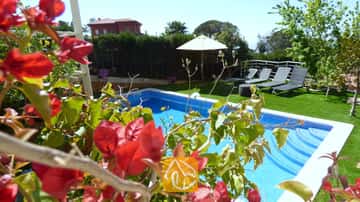 Der private Pool der Villa Noa, eingerahmt von roten Bougainvillea-Blüten, mit Sonnenliegen und einem weißen Sonnenschirm auf dem Rasen im Hintergrund.