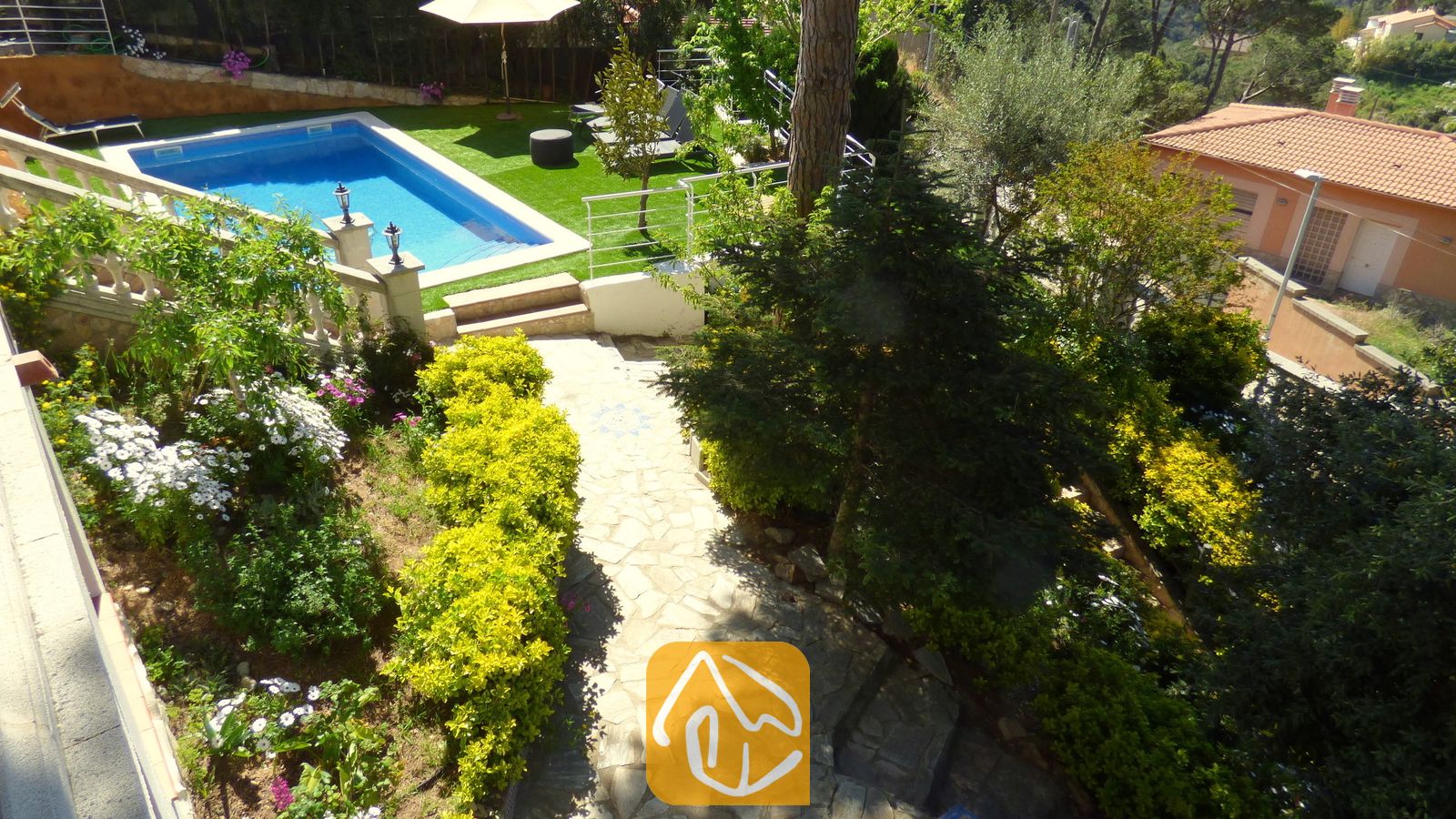 Ansicht von Villa Noa aus der Vogelperspektive mit einem steinernen Gartenweg, üppig grünen Büschen, blühenden Pflanzen und einem rechteckigen Swimmingpool, der von Kunstrasen umgeben ist.
