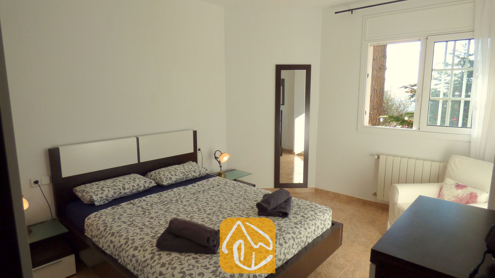 Schlafzimmer mit einem Doppelbett und gemusterter grauer Bettwäsche, zwei zusammengerollten dunklen Handtüchern, einer Nachttischlampe, einem Ganzkörperspiegel und einem Fenster mit Blick ins Grüne.