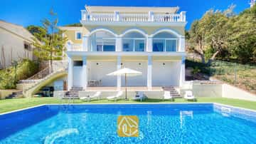 Außenbereich der Villa Sunrise mit einem rechteckigen, blau gefliesten Swimmingpool, weißen Sonnenliegen und einer gelben, mehrstöckigen Villa mit Bogen-Glastüren unter klarem Himmel.