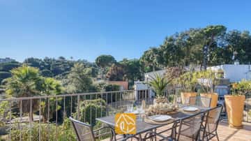 Sonnige Terrasse der Casa AdoRa mit einem Metalldesserttisch, der mit weißen Tellern und Gläsern gedeckt ist, und Blick auf grüne Bäume unter blauen Himmel.