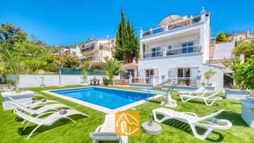 Außenansicht der Villa Maribel mit einem rechteckigen Swimmingpool, Sprungbrett und weißen Sonnenliegen auf grünem Kunstrasen vor einem leuchtend blauen Himmel.