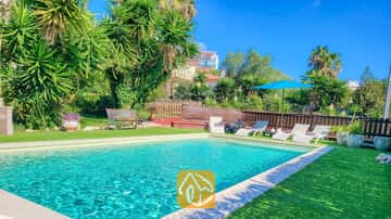 Der Außenpool der Villa Summertime, umgeben von einer grünen Wiese, Sonnenliegen, einem blauen Sonnenschirm und tropischen Palmen unter einem blauen, wolkenlosen Himmel