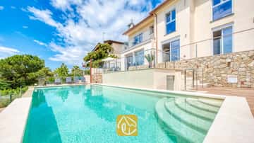 Großer Außenpool mit geschwungener Treppe in der Villa BellaVista. Das mit Naturstein verkleidete Äußere besticht durch Terrassen mit Glasgeländern, Holzterrassen und graue Sonnenliegen, die von einer grünen Baumkulisse umgeben sind.
