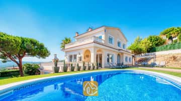 Privater Swimmingpool im Vordergrund mit der Villa Ibiza, einem zweistöckigen, cremefarbenen Haus, grünem Rasen und Kakteen in Töpfen unter einem strahlend blauen Himmel.