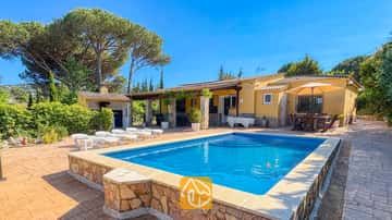 Privater Pool und die gelbe Außenfassade der Villa Iako, mit Steinterrasse, Sonnenliegen und einem Essbereich im Freien unter einem Sonnenschirm vor einem strahlend blauen Himmel.