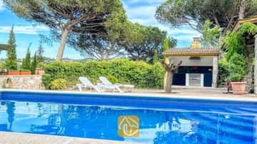 Privater Pool mit blauem Fliesenrand und weißen Sonnenliegen auf der Terrasse der Villa Iako, mit einem Grillhaus im Freien und hohen Pinien im Hintergrund.