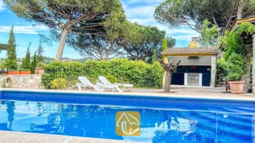 Privater Pool mit blauem Fliesenrand und weißen Sonnenliegen auf der Terrasse der Villa Iako, mit einem Grillhaus im Freien und hohen Pinien im Hintergrund.