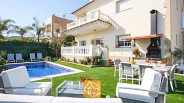 Außenansicht der Villa Mercedes Fenals mit Swimmingpool, Rasenfläche, weißen Gartenmöbeln und einem gemauerten Außen-Grill vor der cremefarbenen Fassade.